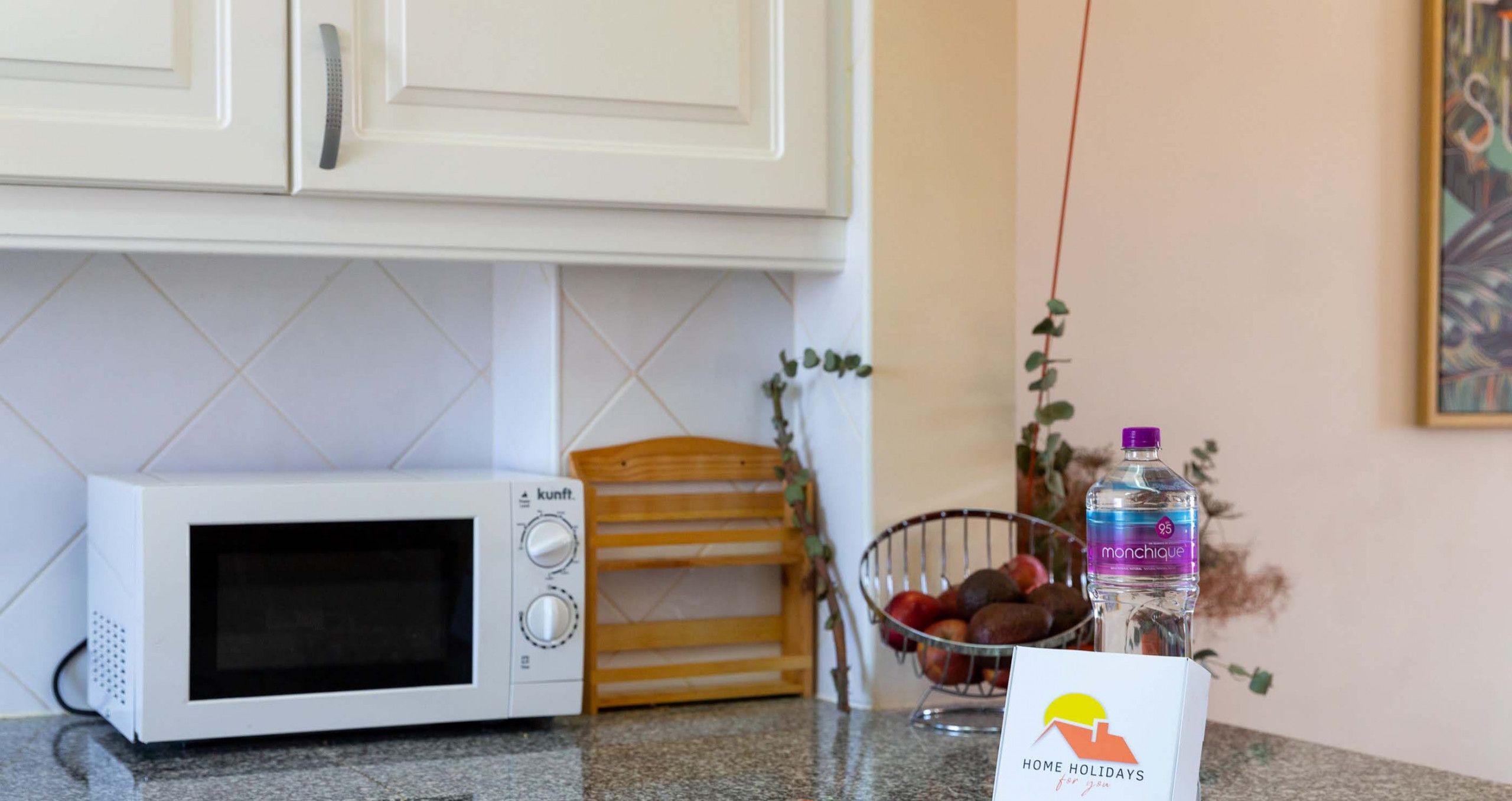 A kitchen with a microwave, a fruit bowl, a water bottle, and a box labeled "HOME HOLIDAYS."