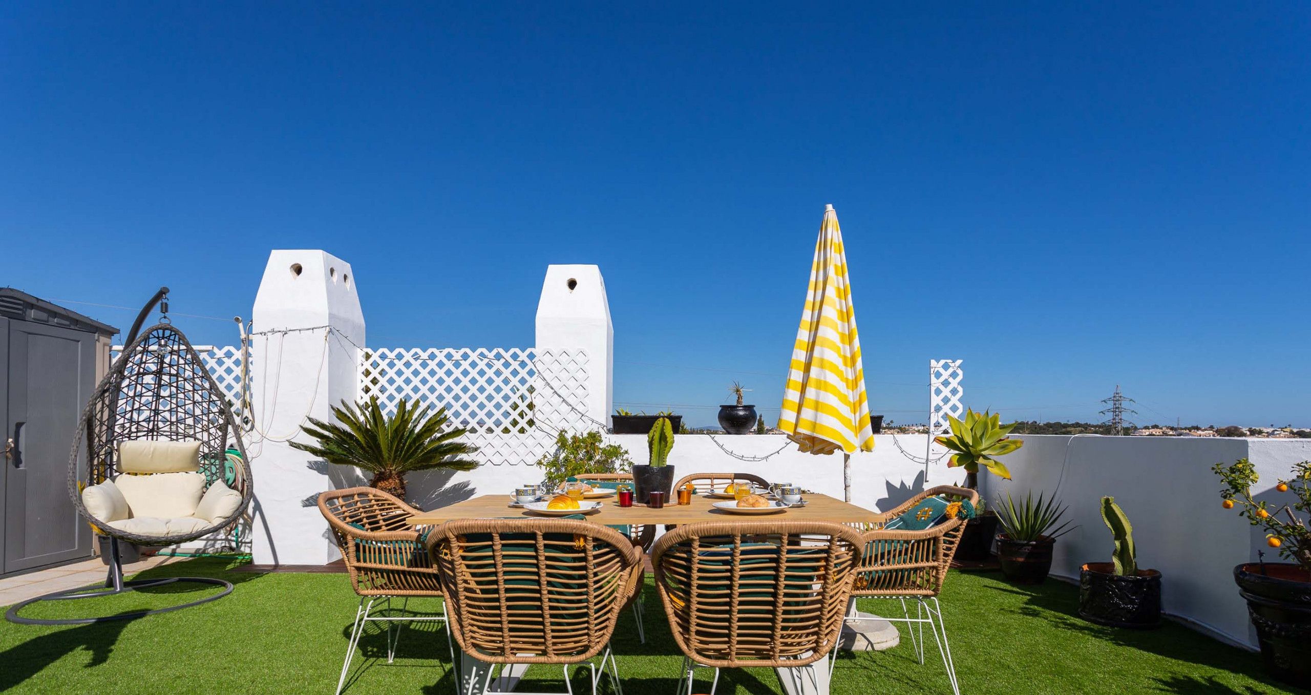 Une terrasse avec une table et des chaises, entourée de plantes en pot et d’une clôture blanche.