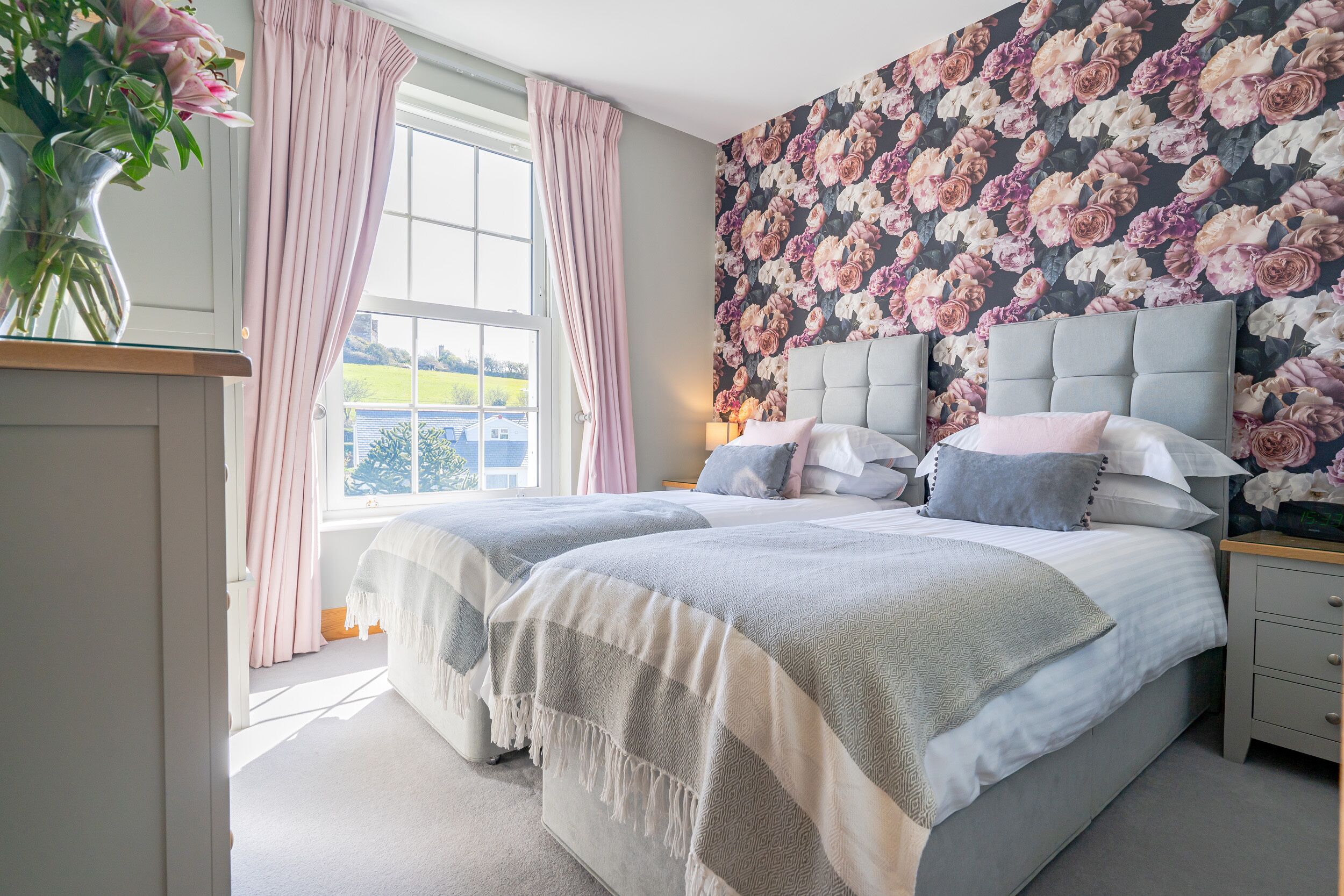 A bedroom with two beds, a floral wallpaper, and a large window with curtains.