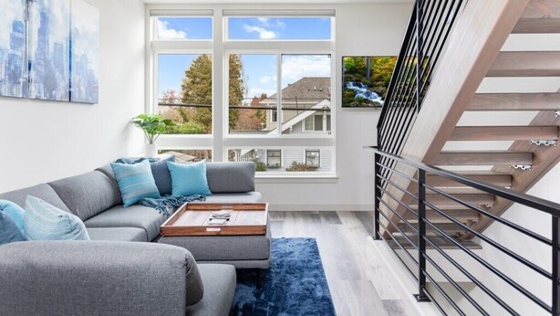 A modern living room with a grey sofa, blue cushions, a wooden coffee table, and a blue rug. There are large windows offering a view of the outside.