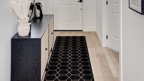 Un couloir avec une porte blanche, un tapis à motifs noir et blanc, un meuble noir orné d’un vase et un luminaire suspendu.