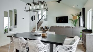 Un espace repas moderne avec une table en bois foncé et des chaises en osier blanches, un escalier offrant une vue sur l’étage supérieur, et un canapé gris en arrière-plan.