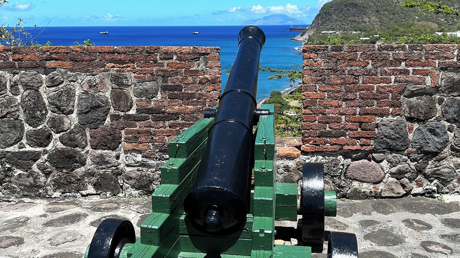 A cannon positioned between stone walls with a view of the ocean and ships in the distance.