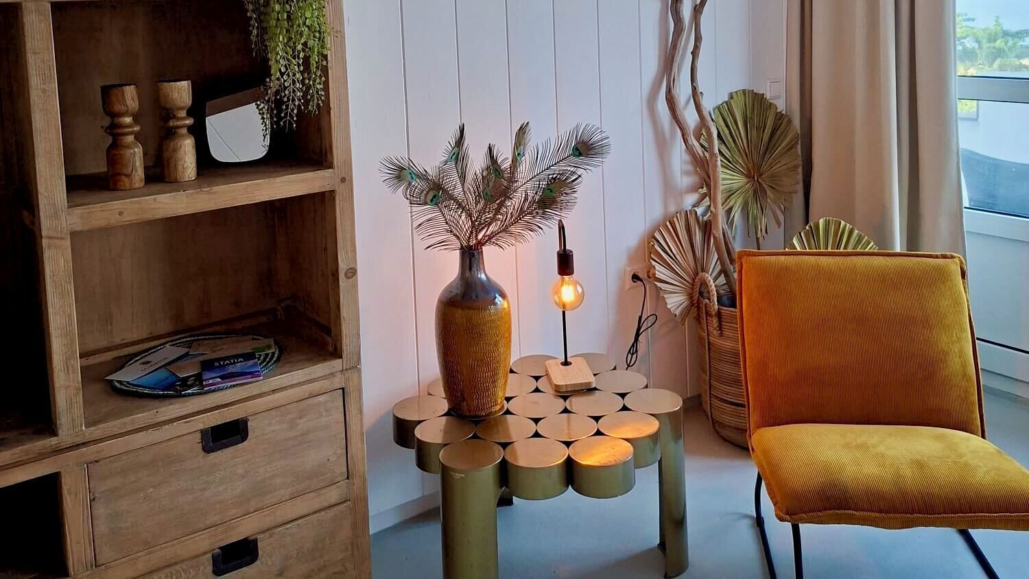 Ett rum med en bokhylla i trä, en gul stol, ett dekorativt bord med en vas och påfågelfjädrar och en växt.