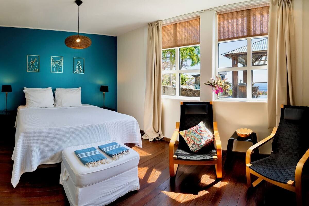 A bedroom with a bed, a chair, a small table, a window with curtains, and artwork on the wall.
