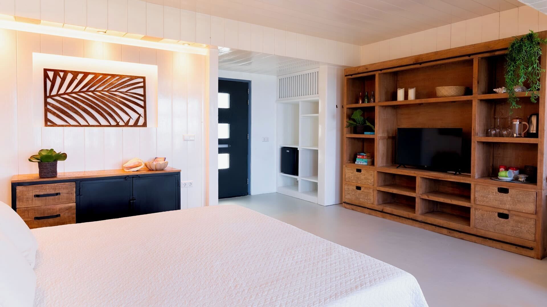 A bedroom with a bed, a wooden TV stand with shelves, a wall-mounted decorative panel, and a door leading to another room.