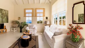 A spacious living room with a white sofa, a coffee table, and a TV. There are two windows with bamboo shades, a large painting, and a mirror on the wall.