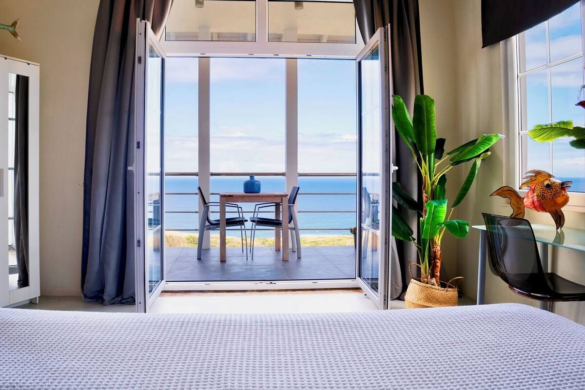 A room with a bed, a table with chairs, a view of the sea, and a balcony with a table and chairs.