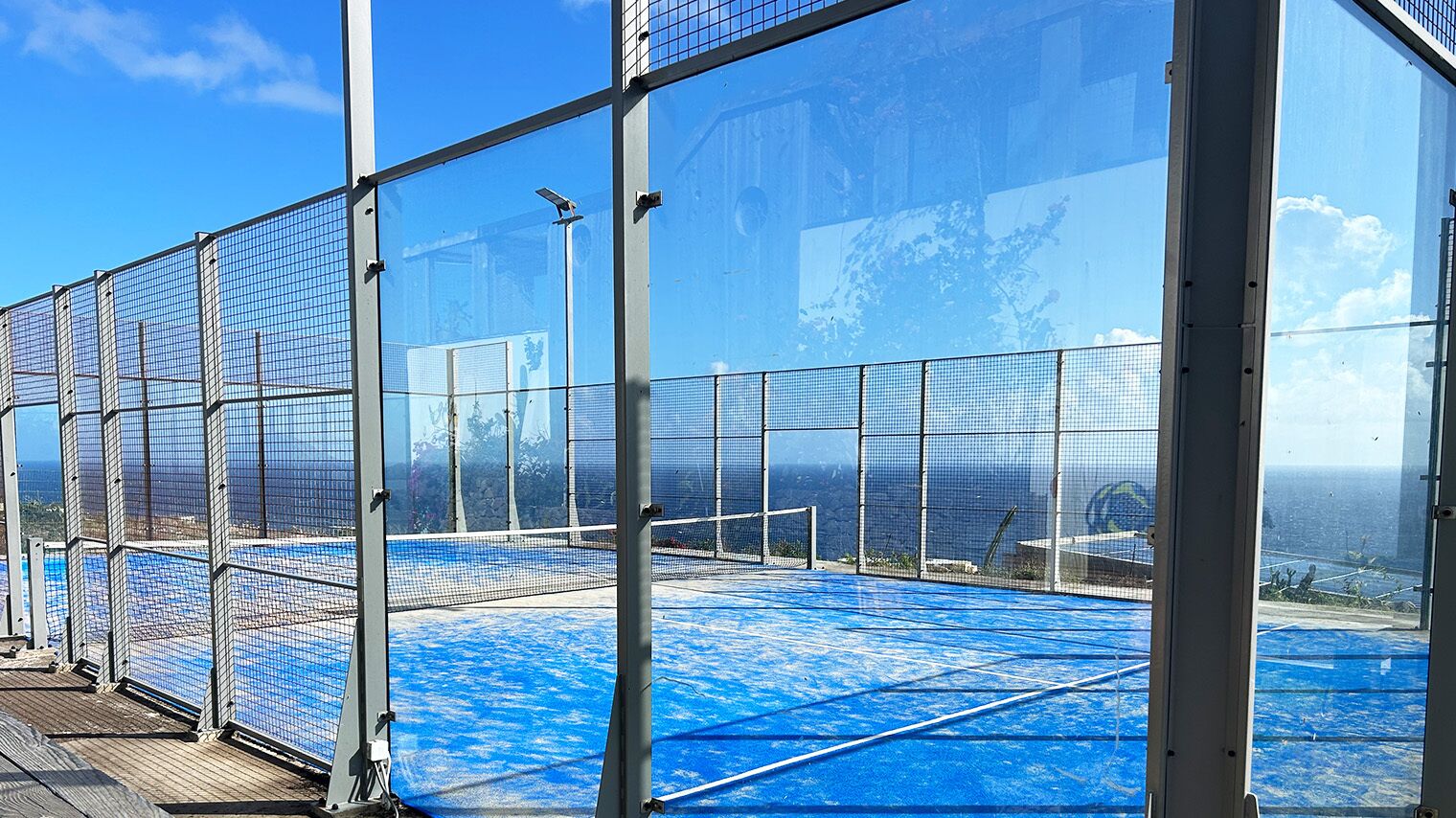 A covered tennis court with netted walls and a blue floor, enclosed by a metal fence.