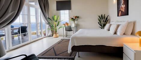 Une chambre à coucher avec un lit, une table de chevet, une lampe, des plantes en pot et une vue dégagée sur l’extérieur grâce à de grandes fenêtres.