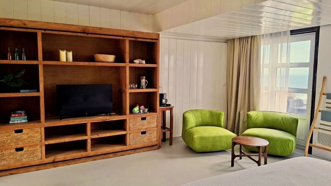 Een kamer met een houten entertainmentsunit, twee groene fauteuils, een kleine tafel en een groot raam met gordijnen.