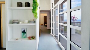 A narrow hallway with white shelving, a hanging plant, and a view of the sea through large windows. - Whale Tails Island Suites & Villas (Oranjestad)