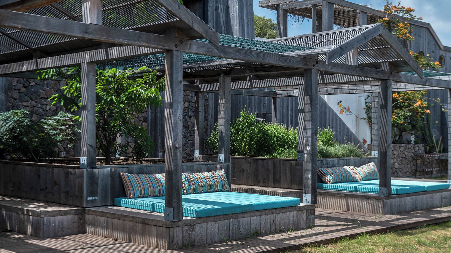 A covered outdoor seating area with wooden benches and cushions, surrounded by greenery and a stone wall.