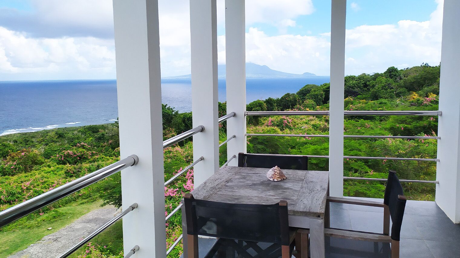 A balcony with a table and chairs, overlooking a coastal landscape with greenery and a view of the ocean.