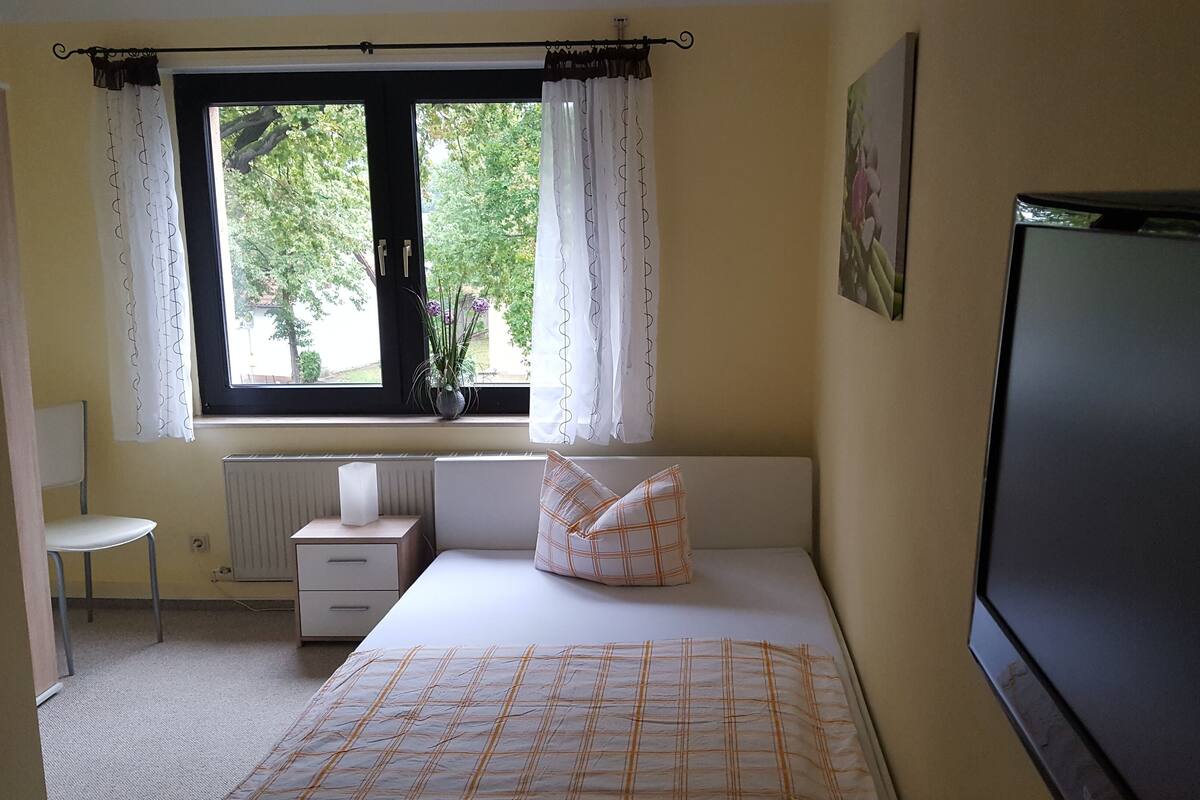 Une chambre à coucher avec un lit, une chaise, un radiateur, une fenêtre avec des rideaux et un téléviseur.