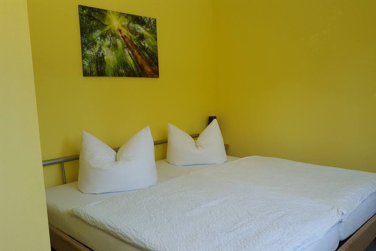 Un lit avec une literie et des oreillers blancs, une tête de lit en bois et un tableau représentant un arbre accroché au mur.