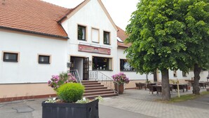Een gebouw met een witte gevel, een rode pannendak en een trap naar de ingang. Er staan buiten tafels en stoelen, en rechts een grote boom.