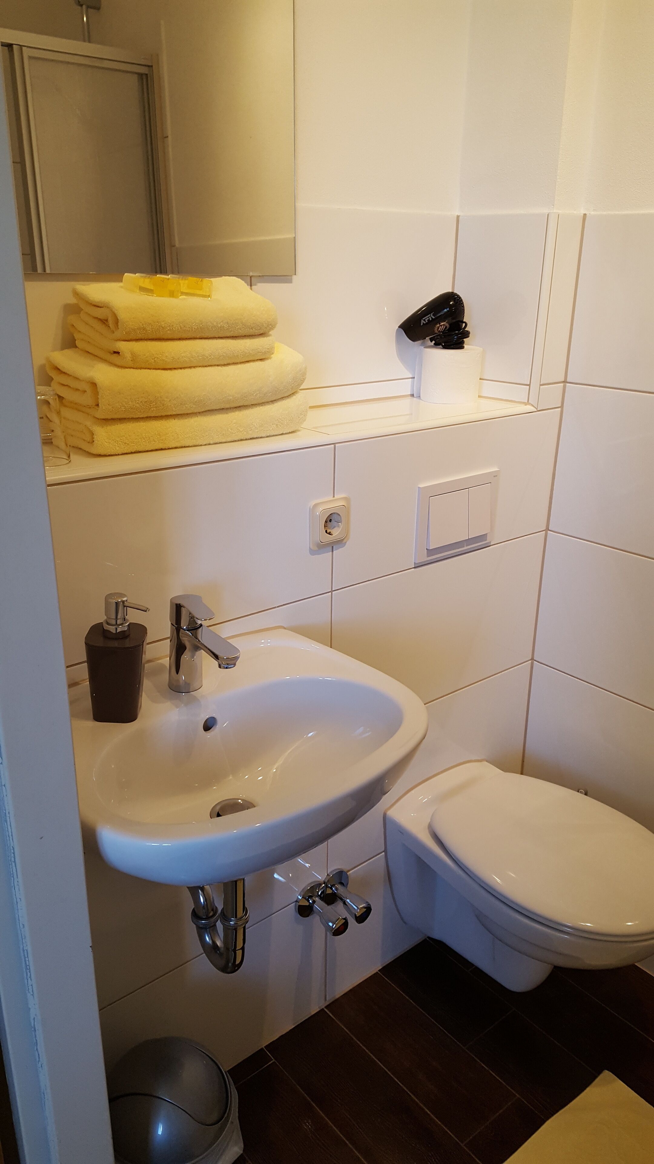 Ein Badezimmer mit Waschbecken, Toilette und Spiegel. Handtücher liegen auf einem Regal und ein Föhn ist an der Wand befestigt.