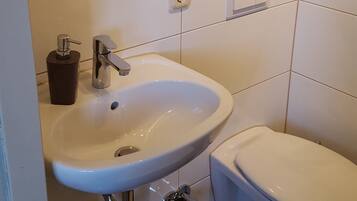 Ein Badezimmer mit Waschbecken, Toilette und Spiegel. Handtücher liegen auf einem Regal und ein Föhn ist an der Wand befestigt.