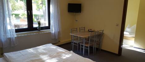 Une pièce avec un lit, une table entourée de chaises, un téléviseur fixé au mur et une fenêtre avec des rideaux.