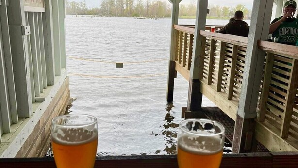 Ein überdachter Außenbereich mit Sitzgelegenheit und zwei Biergläsern auf Untersetzern, mit Blick auf ein Gewässer.