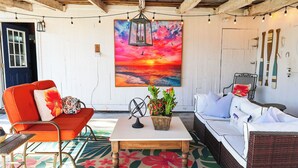 Ein gemütliches Zimmer mit weißer Wand, einem farbenfrohen Sonnenuntergangsbild, einem Korbssessel, einem roten Sessel und einem kleinen Tisch mit einem Korb.