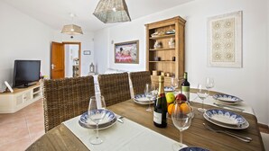 Comedor con una mesa de madera dispuesta para cuatro personas, rodeada de sillas de mimbre. A la izquierda, hay un televisor sobre un mueble bajo, un aparador de madera con objetos decorativos y un cuadro en la pared.