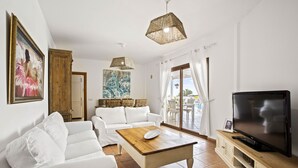 Un salón con un sofá blanco, una mesa de centro de madera, un televisor sobre un mueble de madera y un amplio ventanal con vista al mar.
