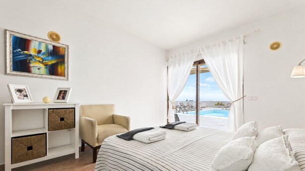 Un dormitorio con cama, una silla, una estantería con cajas de almacenamiento, un cuadro y vista a una piscina.