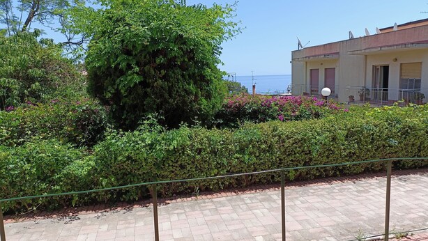 A paved pathway with a metal fence, lush greenery, and a building with a balcony overlooking the sea.