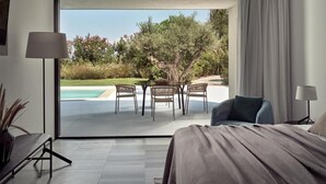 Una habitació d'hotel moderna amb un gran finestral corredís que dona a una zona de piscina, un llit amb llençols grisos, un sofà blau i una taula petita amb cadires.