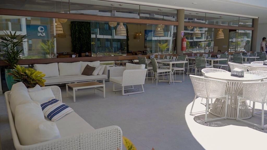A modern indoor lounge area with white wicker furniture, glass walls, and indoor plants.
