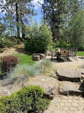 Enjoy dining or socializing in this serene outdoor patio area surrounded by lush landscaping and tall trees.