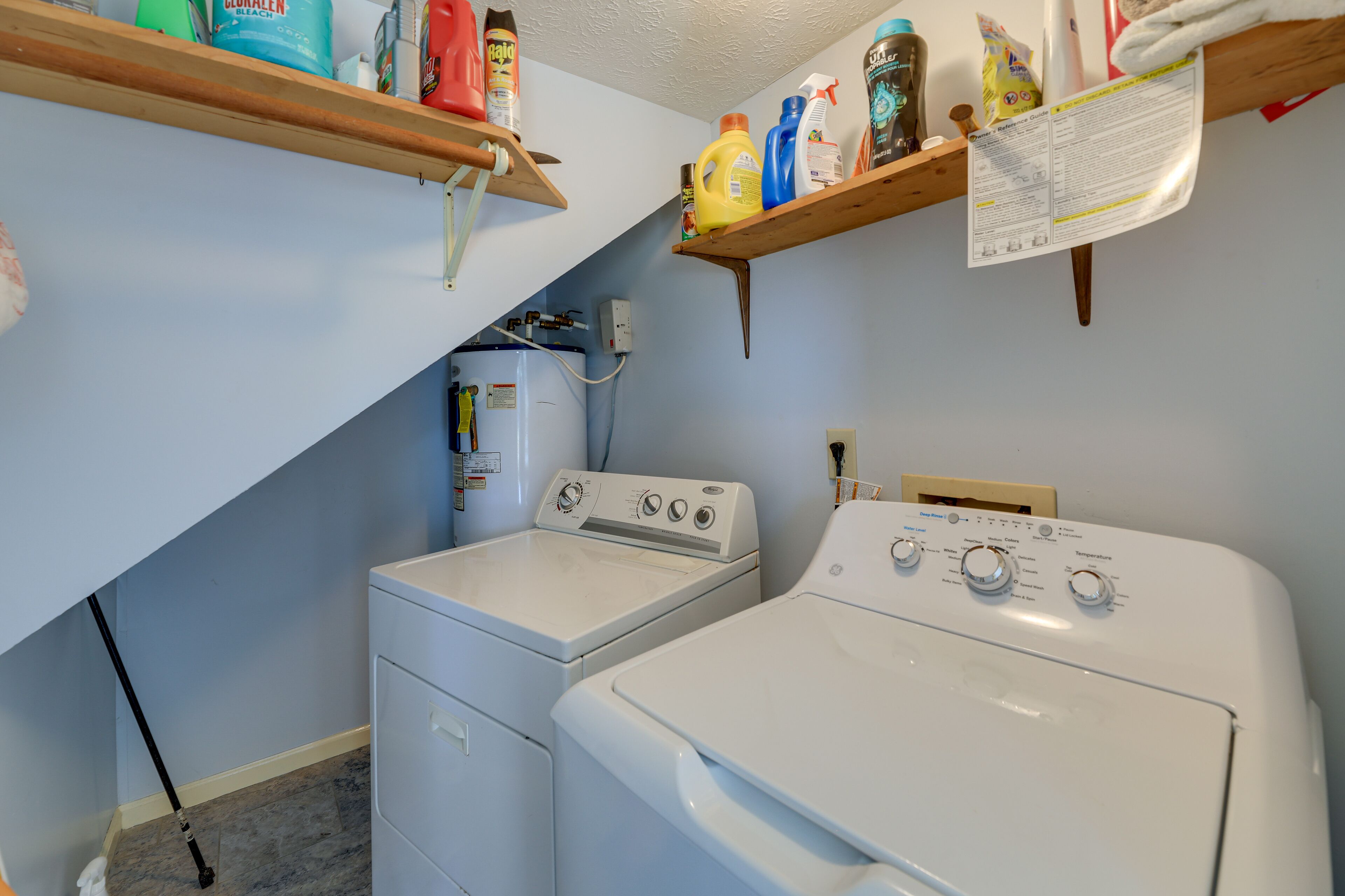 A laundry area with two white washing machines, shelves with cleaning supplies, and a water heater.