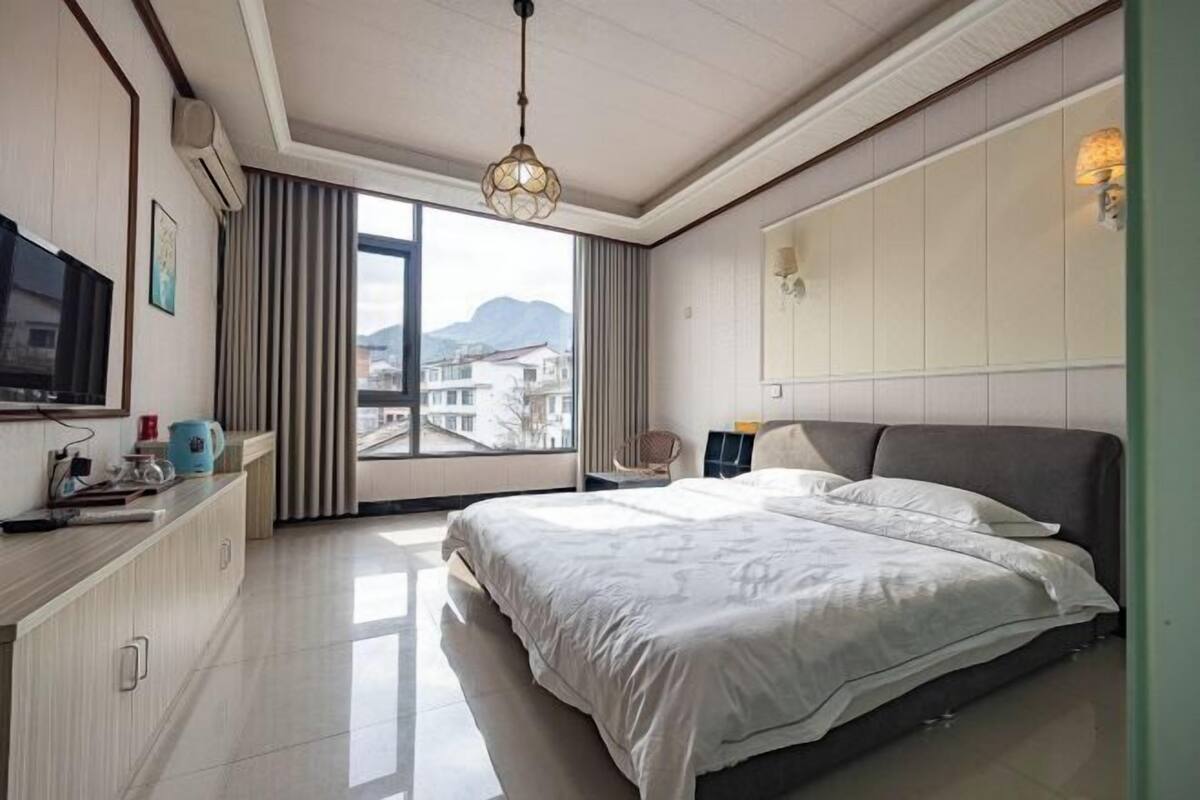 Ein Hotelzimmer mit Bett, Fernseher, Schreibtisch mit Wasserkocher und Blick auf Gebäude durch große Fenster.