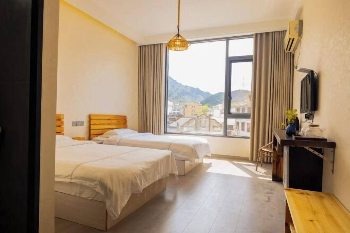 Ein Hotelzimmer mit zwei Betten, einem Schreibtisch, einem Fernseher und einem großen Fenster mit Blick auf Gebäude und Berge.