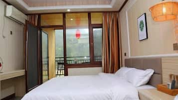 A hotel room with a bed, a bedside table, a window with curtains, an air conditioning unit, and a wall-mounted light fixture.