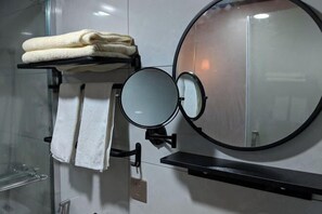 Un baño con cabina de ducha de vidrio, un perchero con toallas dobladas, un espejo redondo y un estante.