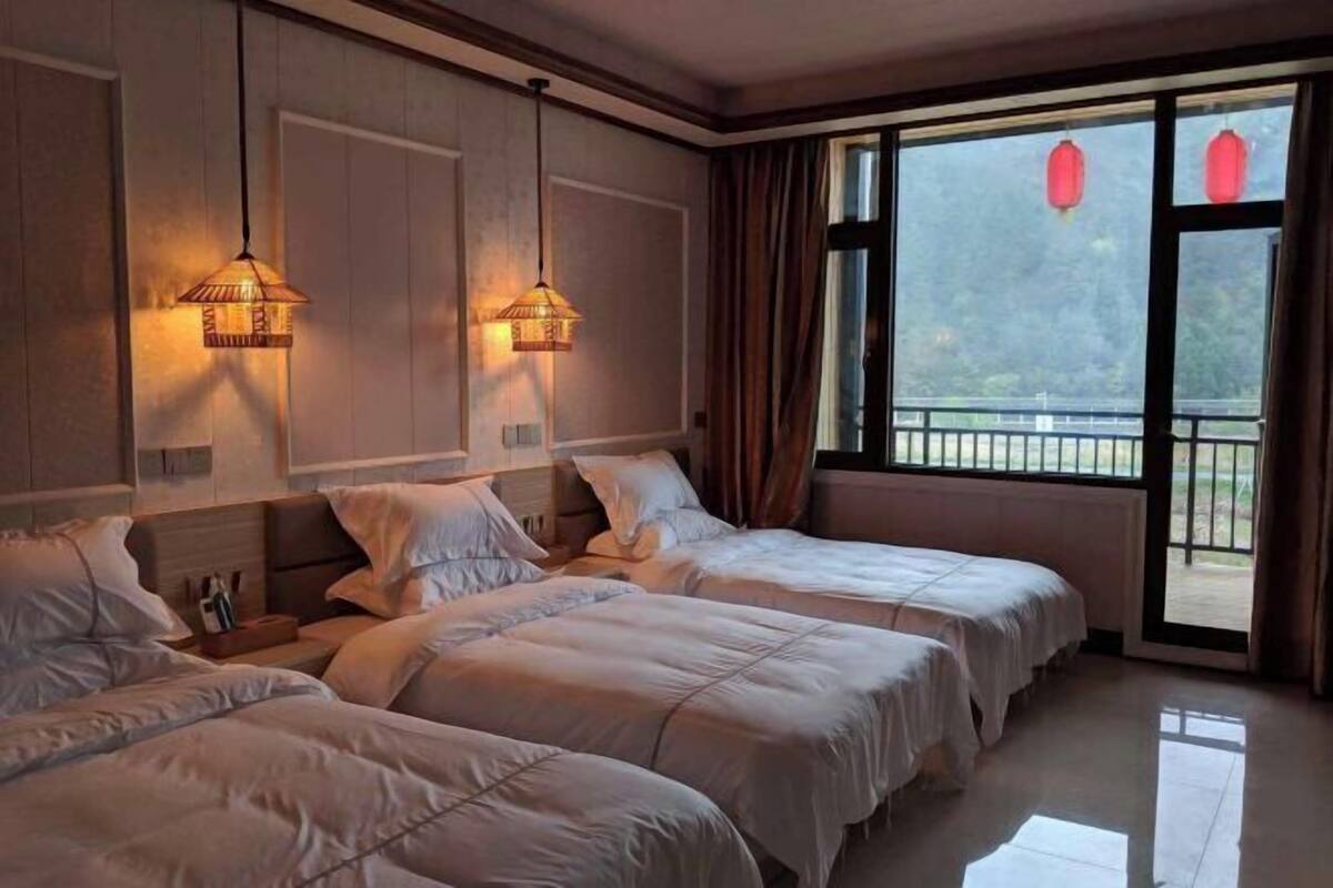Ein Hotelzimmer mit drei Betten, einem großen Fenster mit Vorhängen und Wandleuchten.
