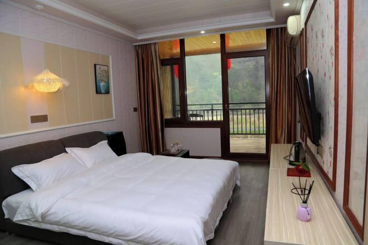 Ein Hotelzimmer mit einem großen Bett, einem Schreibtisch mit einer Vase, einem Fernseher und einem Balkon mit Vorhängen.