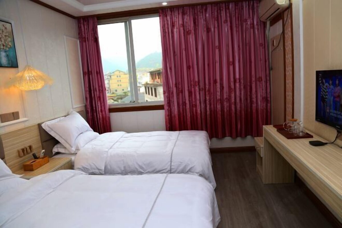 Ein Hotelzimmer mit zwei Betten, einem Fernseher, einem Fenster mit Vorhängen und Blick auf Gebäude und Berge.