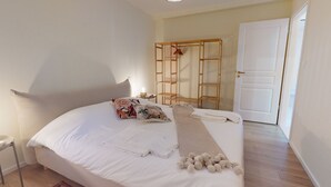 Un lit bien fait, avec des oreillers, une serviette et une couverture. Contre le mur, on aperçoit une étagère en bois et une porte blanche.