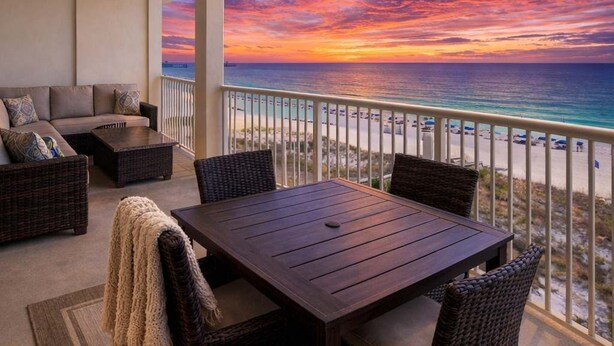 Un balcon avec vue sur l’océan au coucher du soleil, une table à manger, des meubles en osier et un canapé.