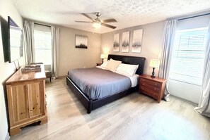 A bedroom with a bed, a nightstand, a dresser, a ceiling fan, and artwork on the wall.