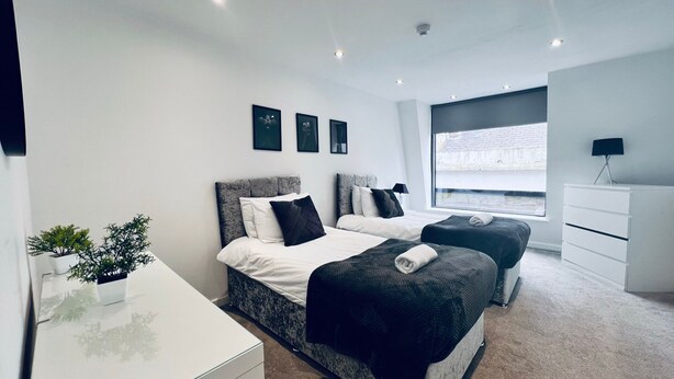A modern bedroom with two beds, a white dresser, a small desk, and a window with a view. - Designer Apartments (Liverpool)