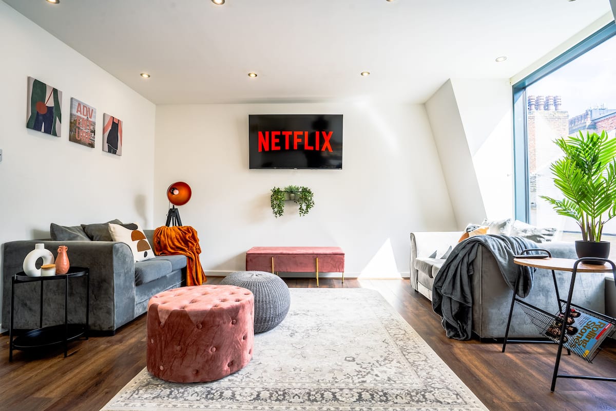 Un salon moderne avec une grande télévision affichant le logo « NETFLIX », un canapé gris, un pouf rouge, un repose-pieds gris et un porte-revues.