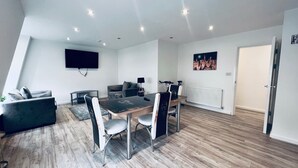 A modern living room with a dining area, a flat-screen TV mounted on the wall, and a cityscape painting. - Designer Apartments (Liverpool)