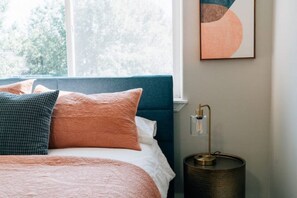 A neatly made bed with a blue headboard, a bedside lamp, and a framed abstract artwork on the wall.