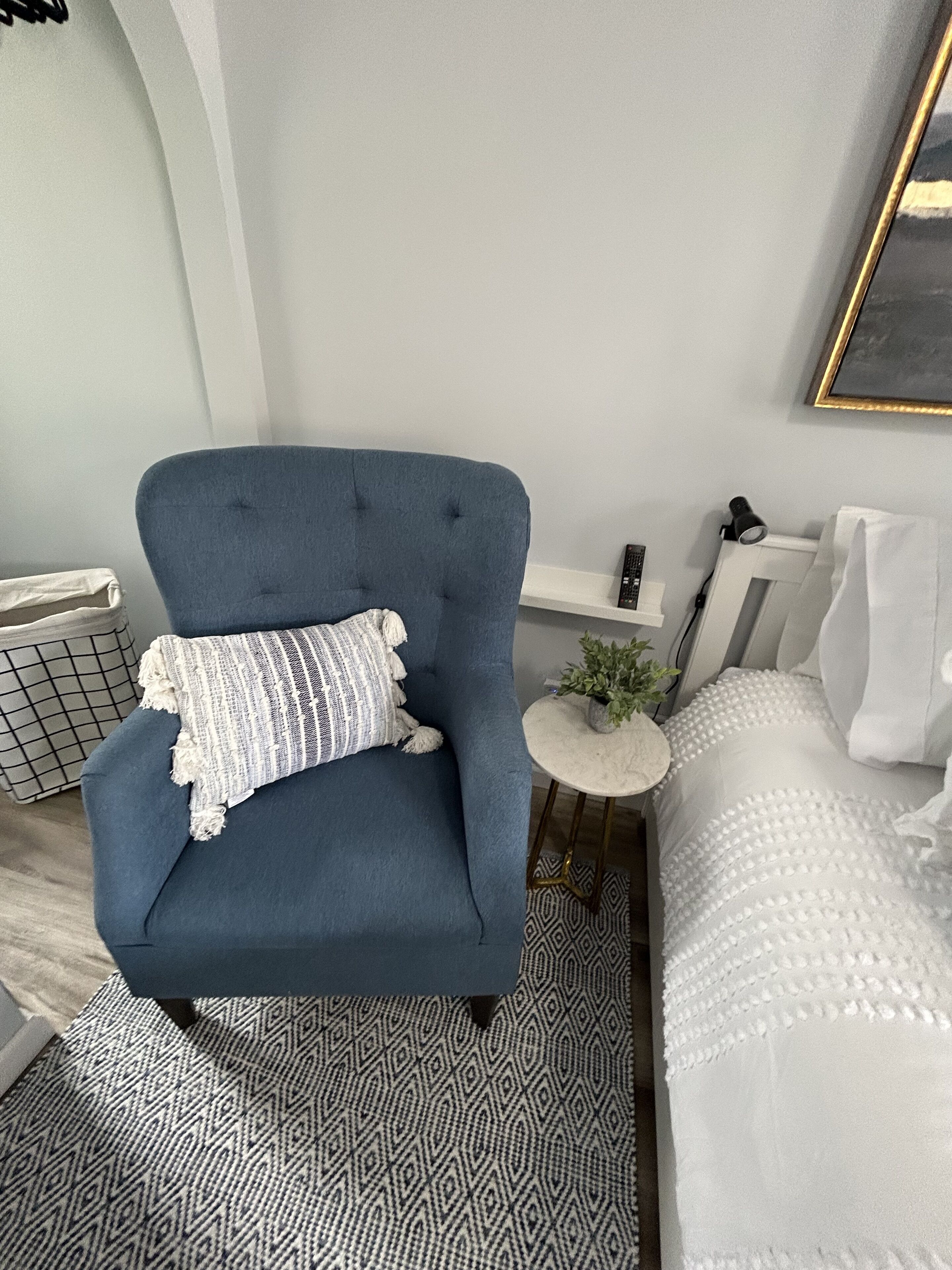 A room with a blue armchair, a white sofa, a small round table with a plant, and a patterned rug.