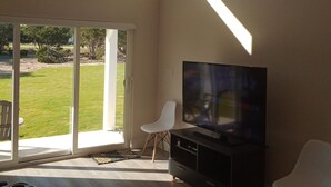 Un ampio soggiorno con una grande TV a schermo piatto, un ventilatore a soffitto e una porta vetrata scorrevole che dà su una zona esterna verdeggiante.